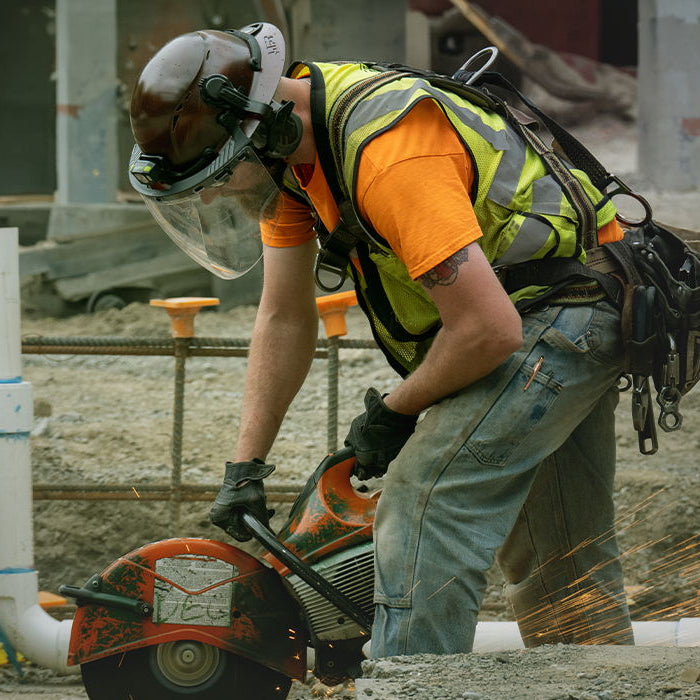 A construction worker wearing a high-visibility safety vest, orange shirt, jeans, and Lift Safety DAX T2 Hard Hat operates a circular saw on a concrete structure, creating bright orange sparks.