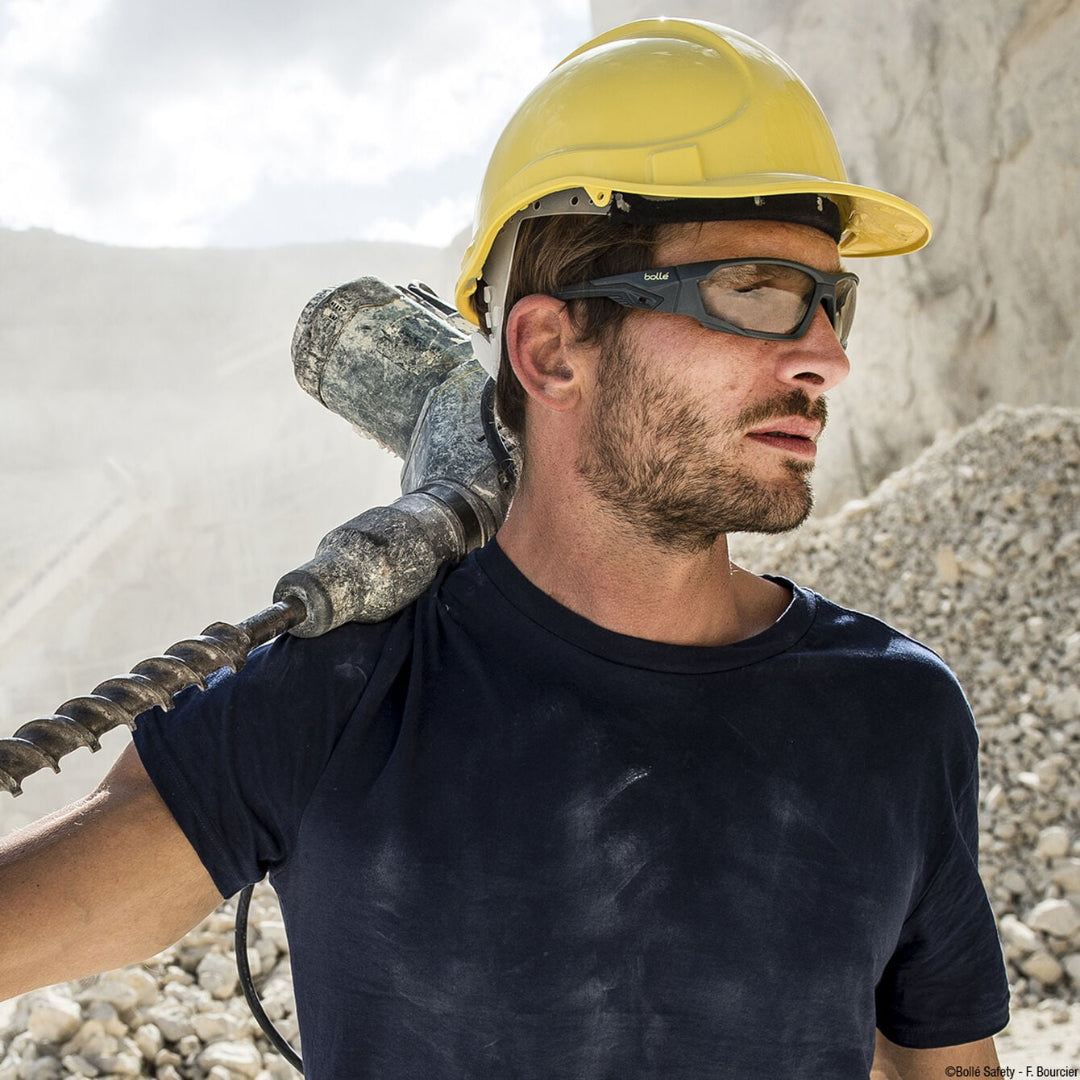 Man wearing a yellow hard hat with Bolle Mercuro Safety Glasses with Copper Lenses holding a power drill against a rocky background