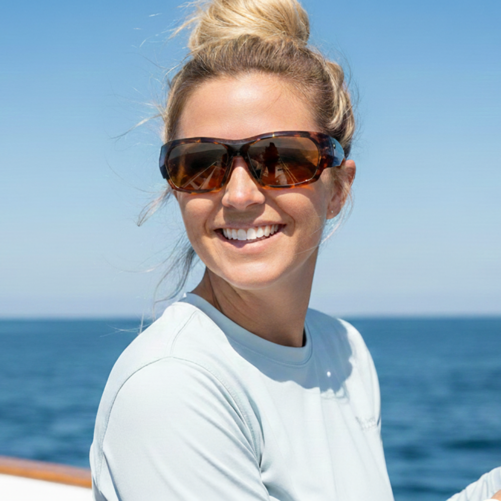 Woman on a sailboat wearing ONOS Nolin Polarized Sunglasses holding a fishing rod with clear blue sky and ocean background